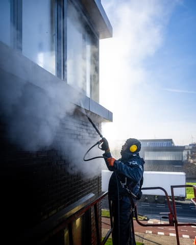 Vakman met gehoorbescherming reinigt gevel met hogedruk bij bedrijfspand in Monster